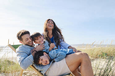 Verspielte Familie umarmt Vater sitzt auf Stuhl am Strand - ASGF03757