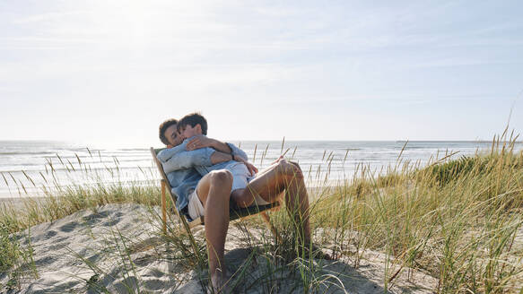 Father embracing son sitting on chair at beach - ASGF03760