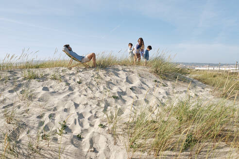 Father relaxing on chair with family playing at beach - ASGF03765