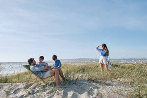 Mutter betrachtet Kinder mit Vater, die sich am Strand an einem sonnigen Tag entspannen - ASGF03767