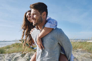 Smiling man piggybacking girlfriend at beach - ASGF03772