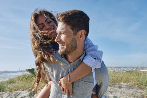 Lächelnder Mann nimmt seine Freundin an einem sonnigen Tag am Strand huckepack - ASGF03773