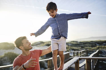 Father supporting son walking on boardwalk railing with arms outstretched at beach - ASGF03778