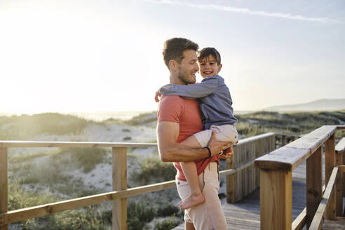 Father carrying son and walking on boardwalk at beach - ASGF03782