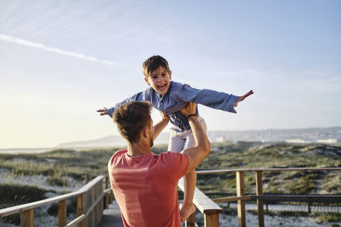 Father carrying son with arms outstretched on boardwalk at beach - ASGF03783