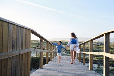 Mother and son holding hands walking on boardwalk at beach - ASGF03788