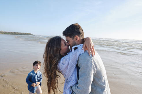 Junge schaut auf romantische Eltern, die sich am Strand küssen - ASGF03807