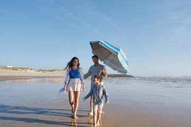 Parents holding sunshade walking with son at beach - ASGF03823