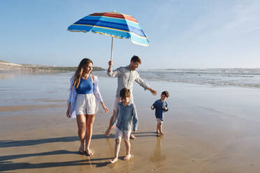 Father holding sunshade walking with family at beach on sunny day - ASGF03826