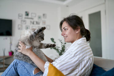 Glückliche Frau streichelt Katze auf Sofa zu Hause - ANNF00332