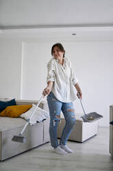 Smiling woman standing with broom in living room at home - ANNF00334