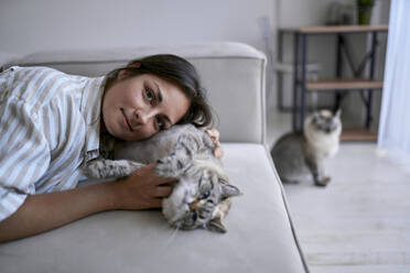 Smiling woman lying with cat on sofa in living room at home - ANNF00336