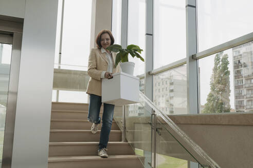 Ältere Geschäftsfrau trägt Kiste mit Topfpflanze auf Treppe in Büro - OSF01710