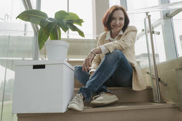 Lächelnde ältere Geschäftsfrau, die neben einer Kiste und einer Topfpflanze auf einer Treppe im Büro sitzt - OSF01713