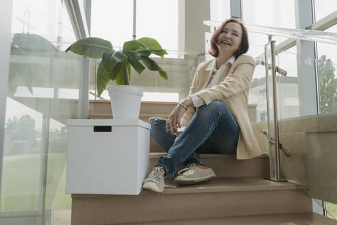 Glückliche ältere Geschäftsfrau, die neben einer Kiste und einer Topfpflanze auf einer Treppe im Büro sitzt - OSF01714