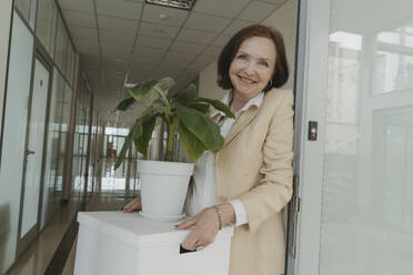 Lächelnde ältere Geschäftsfrau, die eine Schachtel mit einer Topfpflanze im Korridor eines Büros hält - OSF01715