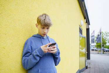 Junge mit Smartphone vor einer gelben Wand stehend - NJAF00348