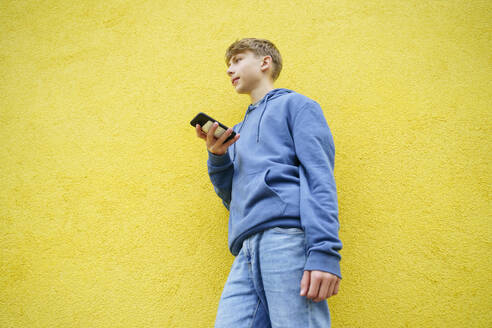 Junge mit Smartphone vor einer gelben Wand stehend - NJAF00350