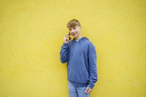 Lächelnder Junge, der vor einer gelben Wand mit seinem Smartphone spricht - NJAF00351