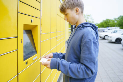 Junge mit Fahrkarte vor einem Paketkasten stehend - NJAF00357