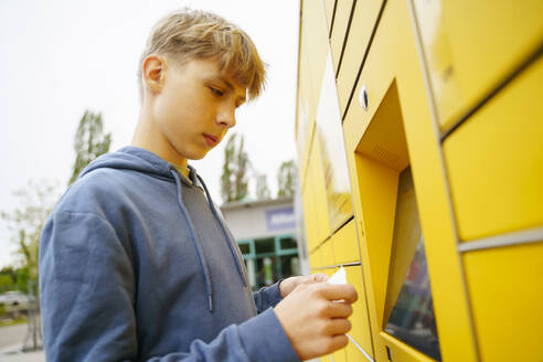 Boy with ticket standing in front of yellow parcel locker - NJAF00359