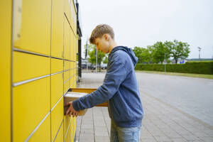 Blonder Junge nimmt Paket aus Briefkasten am Fußweg - NJAF00361