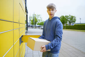 Junge mit Paket neben gelbem Paketkasten stehend - NJAF00362