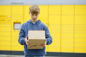 Junge, der ein Paket vor einem gelben Paketkasten prüft - NJAF00363