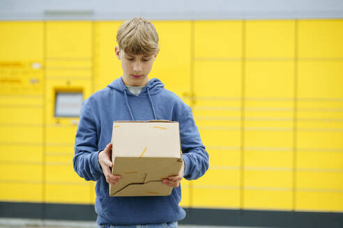 Junge, der ein Paket vor einem gelben Paketkasten prüft - NJAF00363