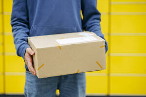 Junge mit Paket vor einem gelben Paketkasten stehend - NJAF00364