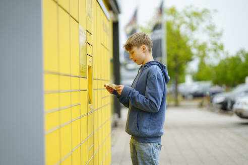 Blond boy holding ticket standing in front of parcel locker at footpath - NJAF00366