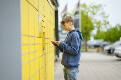 Blonder Junge mit Fahrkarte vor einem Paketkasten am Fußweg stehend - NJAF00366