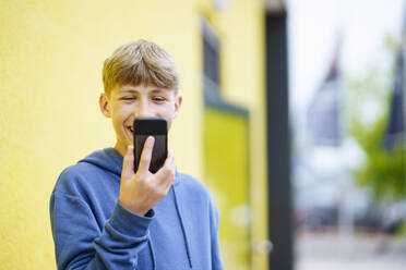 Glücklicher blonder Junge, der vor einer gelben Wand ein Videogespräch führt - NJAF00369