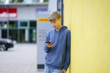 Blonder Junge lehnt an einer gelben Wand und benutzt ein Mobiltelefon - NJAF00371