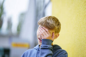 Blonder Junge verdeckt sein Gesicht mit der Hand vor einer gelben Wand - NJAF00372