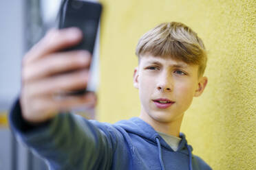 Blond boy taking selfie through smart phone in front of yellow wall - NJAF00373