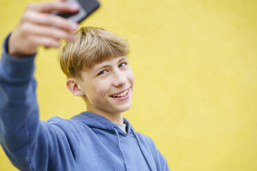 Fröhlicher blonder Junge macht ein Selfie mit seinem Smartphone vor einem leuchtend gelben Hintergrund - NJAF00374