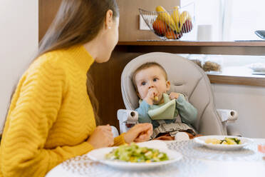 Mother looking at baby boy eating solid food on high chair at home - NDEF00741