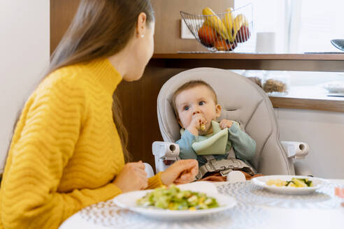 Mother looking at baby boy eating solid food on high chair at home - NDEF00741