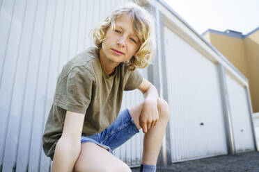 Junge mit blondem Haar hockt vor der Garage - NJAF00378