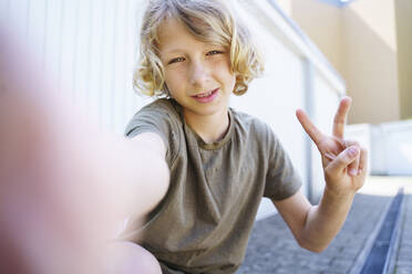 Junge mit blondem Haar zeigt Friedenszeichen vor Garage - NJAF00379
