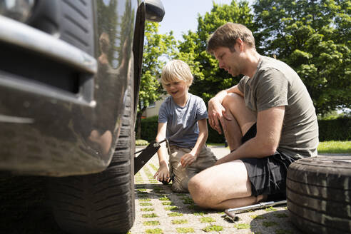 Smiling son sitting by father changing car tire in yard - NJAF00385