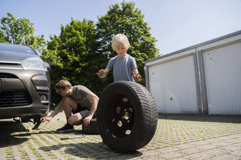 Boy helping father changing car tire in yard - NJAF00391