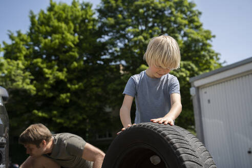 Junge rollt Reifen mit Vater repariert Auto im Hintergrund - NJAF00392