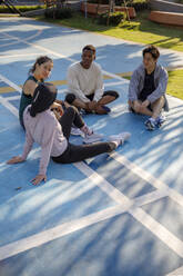 Friends talking to each other sitting on floor in sports court - IKF00884