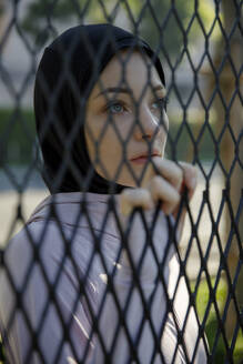 Thoughtful woman wearing hijab holding chainlink fence - IKF00886