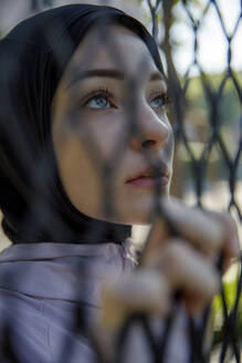 Frau mit Hidschab, die hinter einem Maschendrahtzaun nachdenkt - IKF00887