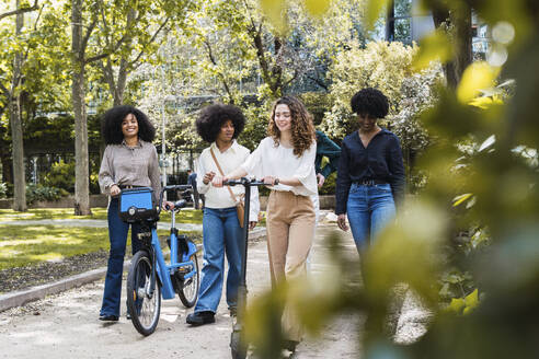 Glückliche junge Freunde, die zusammen mit einem Elektrofahrrad und einem Tretroller im Park spazieren gehen - PNAF05351