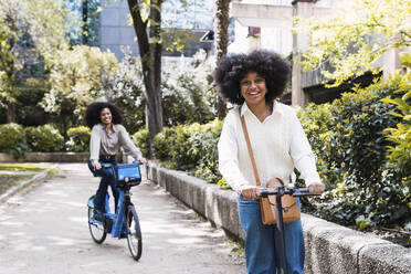 Happy young friends riding electric push scooter and bicycle together in park - PNAF05355