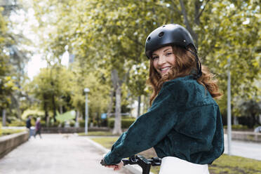 Lächelnde junge Frau mit Elektro-Scooter im Park stehend - PNAF05376